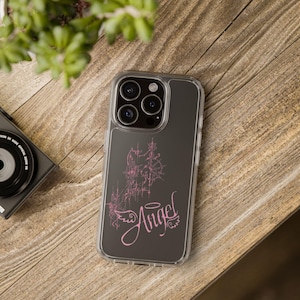 Puede incluir: Funda de teléfono transparente con una parte trasera gris oscuro. Presenta un diseño gráfico rosa y la palabra "Angel" en cursiva. La funda está sobre una superficie de madera, con una cámara y vegetación en el fondo.