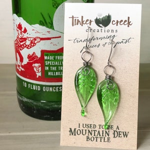 May include: Green glass leaf earrings with silver wire accents. The earrings are hanging from a card that reads "Tinkercreek creations transforming pieces of the past" and "I used to be a Mountain Dew bottle".
