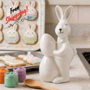 May include: A white ceramic bunny figurine holding an egg. The bunny has closed eyes and a black nose and mouth. Easter bunny biscuits are on a tray in the background.