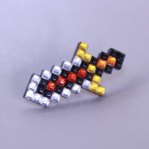 Könnte beinhalten: Ein pixeliger schwertförmiger Ansteckpin mit einem Mosaikdesign. Der Pin weist eine Kombination aus schwarzen, weißen, roten, orangefarbenen und gelben quadratischen Edelsteinen auf. Der Pin ist vor einem hellgrauen Hintergrund platziert.