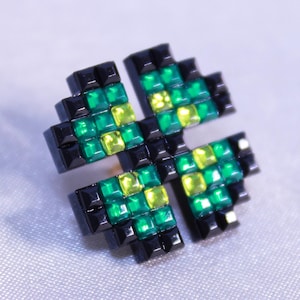 Puede incluir: Broche en forma de trébol de cuatro hojas con diseño pixelado. El broche presenta cristales cuadrados negros, verdes y amarillos. Los cristales están dispuestos para formar las hojas y el contorno del trébol.