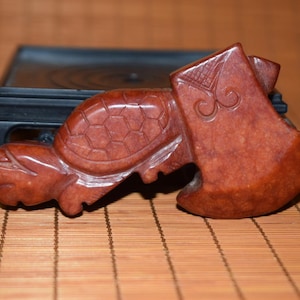 Puede incluir: Escultura de piedra tallada de color marrón rojizo de una tortuga con forma de hacha. La tortuga tiene un patrón de caparazón detallado y descansa sobre un soporte negro. El fondo es una estera de bambú.