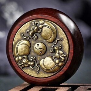 May include: A round, wooden decorative piece with a dark reddish-brown frame. The center features a gold-colored metal design with three mythical creatures surrounding a Yin and Yang symbol. The design is detailed and raised.