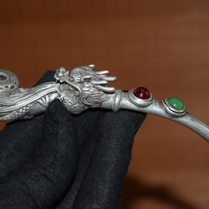 May include: A silver-colored pipe with a dragon head design. The pipe features a bowl with a decorative rim and a stem adorned with a red and a green gemstone. The pipe is held against a dark background.
