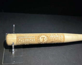 May include: A wooden baseball bat with engraved text. The text reads "2023 AMERICAN LEAGUE CHAMPIONS" and "2023 WORLD SERIES CHAMPIONS" with the Texas Rangers logo. The bat is light brown and displayed on a clear stand.