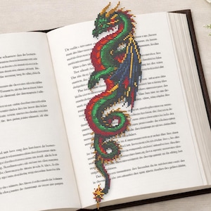 Puede incluir: Un marcapáginas de dragón bordado y colorido se encuentra en un libro abierto. El dragón presenta tonos verdes, rojos y azules con detalles amarillos. También se ve un libro encuadernado en cuero y un ramo de flores blancas.