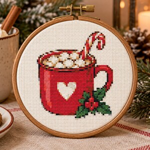 May include: A cross-stitch embroidery piece in a round wooden hoop featuring a red mug filled with white marshmallows and a candy cane. The mug is decorated with a white heart and green holly leaves with red berries at the base on white fabric.