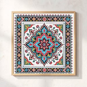 May include: A square cross-stitch embroidery piece framed in light wood, featuring a symmetrical floral mandala pattern in vibrant shades of turquoise, red, yellow, and black on a white background, enclosed by a decorative geometric border.