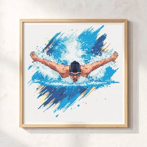 May include: Framed artwork featuring a swimmer in the butterfly stroke. The image uses a pixelated style with shades of blue, white, and tan to depict the swimmer and water. The artwork is in a light wood frame, suitable for sports enthusiasts.