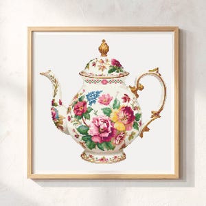 May include: A framed cross-stitch of a floral teapot. The teapot is white with pink, yellow, and blue flowers. It has a gold handle and spout, and a gold finial on the lid. The frame is light brown.