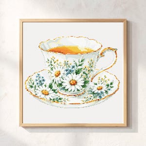 May include: A square framed cross-stitch pattern featuring a white teacup and saucer filled with amber tea. The porcelain is decorated with white daisy flowers, green leaves, and small blue blossoms, all outlined with a delicate gold rim.