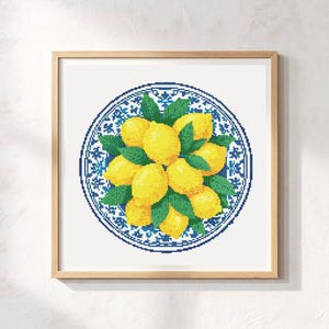 May include: Cross-stitch artwork featuring a plate with a blue and white floral pattern, laden with yellow lemons and green leaves. The piece is framed in a light brown wooden frame, set against a white backdrop.