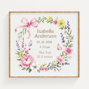 Op de afbeelding: Een ingelijste kruissteek geboorteaankondiging met een bloemenkrans van roze, gele en paarse bloemen, een roze strik en twee vlinders. De tekst luidt Isabella Anderson, 03.10.2026, 3.21 pm, 7lbs 8oz en 20.6 inches in een bruin lettertype.
