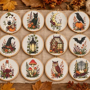 Op de afbeelding: Twaalf ronde houten borduurringen met kruissteekontwerpen van herfst- en Halloween-motieven, waaronder een geest, zwarte kat, maansikkel, raaf, heksenhoed, lantaarn, kas, vleermuis, toverdrankfles, paddenstoelen, uil en een kruiwagen.