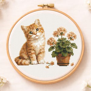 Op de afbeelding: Kruissteekborduurwerk van een rood katje naast een potplant met perzikbloemen. De borduurwerk is geplaatst in een houten borduurring, wat een charmant, handgemaakt ontwerp laat zien.