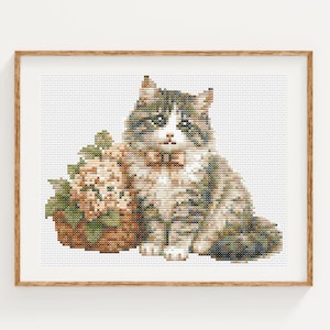 Può includere: Uno schema a punto croce raffigurante un gatto tigrato dal pelo bianco e grigio, che indossa un piccolo papillon, seduto accanto a un cesto intrecciato pieno di fiori rosa chiaro. Il disegno è inserito in una semplice cornice in legno chiaro.