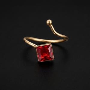 May include: A gold-toned open ring featuring a single square-cut red gemstone. The band is thin and flexible, with a small spherical accent at the open end, while the square red stone is set securely at the opposite end of the bypass design.