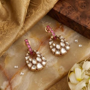 May include: A pair of gold-toned earrings with a teardrop design. The earrings feature a cluster of white stones within an openwork frame. The top portion of the earrings is a pink rectangular bar. The earrings are displayed on a marble surface.