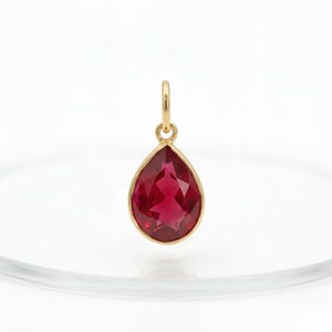 May include: A teardrop-shaped pendant with a faceted, deep red gemstone set in a gold-coloured bezel. The pendant has a small loop at the top for a chain. The gemstone is approximately 2 cm long.