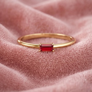 Puede incluir: Un anillo de oro con una piedra preciosa rectangular de color rojo intenso. El anillo está sobre una superficie de terciopelo rosa suave. La piedra preciosa es el punto focal, mostrando un color vibrante y un diseño elegante.