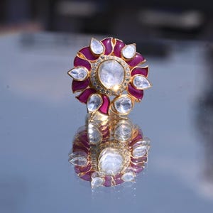 Puede incluir: Un anillo dorado con un diseño floral. El anillo presenta una piedra central grande, clara y redonda, rodeada de piedras claras más pequeñas y pétalos de color rosa intenso. El anillo se refleja en la superficie brillante de abajo.