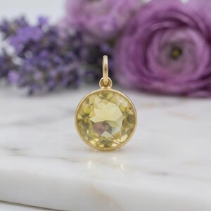 May include: A round, gold-colored pendant with a large, faceted yellow gemstone. The pendant is set against a blurred background of purple flowers and rests on a white marble surface. The gemstone is a light yellow color.