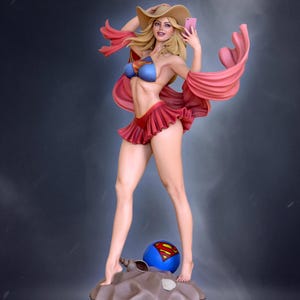 May include: A statue of a woman in a red and blue swimsuit, a tan hat, and a pink scarf, holding a pink phone. A blue ball with a Superman logo is at her feet. The statue is on a sandy base.