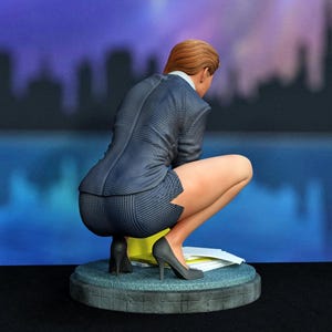 Könnte beinhalten: Eine Figur mit hellbraunem Haar, bekleidet mit einem dunkelblauen, gestreiften Blazer und passendem Rock, hockt auf einem runden grauen Sockel. Die Figur trägt dunkelgraue High Heels und befindet sich neben einem Stapel weißer Papiere.