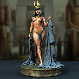 Pode incluir: Uma estatueta de uma figura com cabelo escuro usando um adorno de cabeça dourado, braçadeiras douradas e uma corrente dourada na cintura. A figura está envolta em um tecido azul estampado sobre uma base circular com marcas esculpidas.