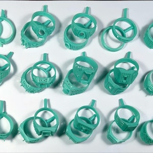 May include: A collection of teal-colored ring molds, each with a unique design. The rings vary in style, some featuring geometric patterns, others with textured surfaces. Each mold has a small handle attached, suggesting they are for jewelry making.