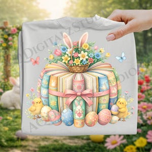 Könnte beinhalten: Ein hellgraues Sweatshirt mit einem farbenfrohen Osterdesign. Das Design zeigt einen Stapel Bücher, einen Blumenkorb, Hasenohren, dekorierte Ostereier und Küken. Eine rosa Schleife umschließt die Bücher. Auch Schmetterlinge sind vorhanden.