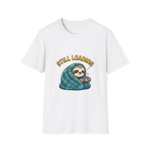 May include: White t-shirt featuring a cartoon sloth wrapped in a blue and green plaid blanket, holding a coffee cup. The text "STILL LOADING" is above the sloth in yellow letters.