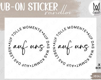 May include: Two circular rub-on stickers featuring black text on a white background. The center reads "auf uns" in a cursive script, surrounded by the circular text "AUF TOLLE MOMENTE AUF DAS WAS KOMMT AUF DAS LEBEN" with small heart icons.
