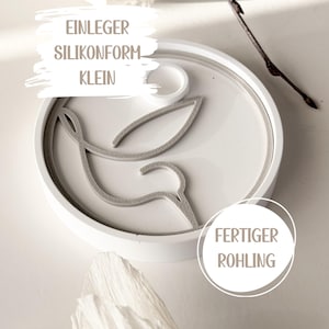 Könnte beinhalten: Eine weiße Silikonform mit einem Taubendesign. Die Form ist rund mit einem grauen Taubenumriss und dem Text "EINLEGER SILIKONFORM KLEIN" und "FERTIGER ROHLING". Ein Zweig und eine Papierblume befinden sich im Hintergrund.