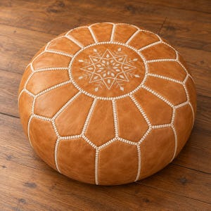 May include: A round, tan leather pouf featuring a central white embroidered snowflake-like geometric pattern. The surface is divided into panels by white stitched seams, creating a structured, segmented appearance across the top and sides.