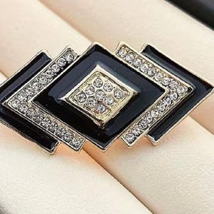 Könnte beinhalten: Geometrische Brosche mit schwarzem Emaille-Finish und goldfarbenen Akzenten. Die Brosche zeigt rautenförmige Elemente, die jeweils mit funkelnden, klaren Kristallen umrandet sind. Die zentrale Raute ist mit kleineren Kristallen gefüllt.