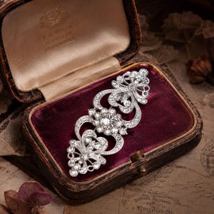 Puede incluir: Un broche de metal plateado con un diseño alargado y simétrico, adornado con numerosas piedras transparentes brillantes. La pieza presenta intrincados motivos de volutas y flores, y se encuentra dentro de un joyero forrado de terciopelo.