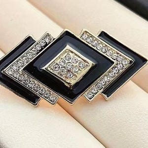 May include: A geometric brooch with a black enamel finish and gold-tone accents. The brooch features a series of diamond-shaped elements, each embellished with sparkling clear crystals. The central diamond is filled with smaller crystals.