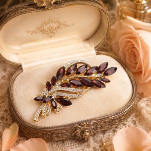 Op de afbeelding: Een goudkleurige broche in de vorm van een bladerige tak, voorzien van dieprode, granaatkleurige stenen in marquise-slijpvorm en kleine heldere witte kristallen langs de metalen stelen. Het sieraad ligt in een ovaal, bewerkt metalen doosje.