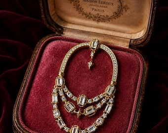 May include: A gold brooch featuring an oval frame encrusted with small clear stones, with three dangling chains adorned with baguette and round clear stones. A small star-shaped charm hangs from the top center of the oval frame.