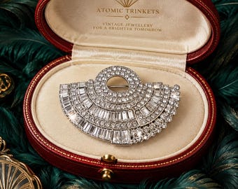 May include: A silver-toned fan-shaped brooch encrusted with clear, sparkling rhinestones in a mix of baguette and round cuts. The brooch is displayed inside an oval, deep red velvet-lined jewelry box with the text ATOMIC TRINKETS VINTAGE JEWELLERY.
