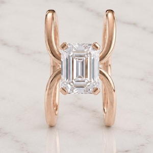 May include: A rose gold ring featuring a central emerald-cut clear gemstone held by four prongs. The band consists of two curved, parallel metal segments that extend vertically from the stone, creating an open, architectural design.