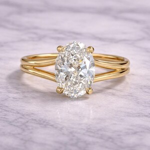 Könnte beinhalten: Ein goldener Ring mit einem großen, oval geschliffenen Diamanten. Das Goldband ist in zwei parallele Stränge geteilt. Der Diamant funkelt im Licht. Das Design ist elegant und ansprechend.