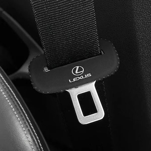 Puede incluir: Una funda de cinturón de seguridad negra con el logotipo y el texto de Lexus en blanco. La funda es rectangular con bordes redondeados y está colocada sobre una correa de cinturón negra con una hebilla de metal plateado visible debajo.
