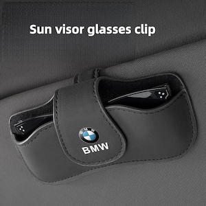 May include: A black sun visor glasses clip featuring the BMW logo and text on the front flap. The holder is attached to a dark gray fabric surface and contains a pair of black glasses inside the storage compartment.
