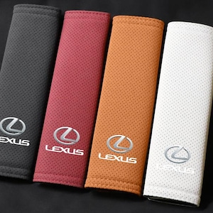 Puede incluir: Cuatro fundas rectangulares para cinturón de seguridad en negro, rojo oscuro, tostado y blanco. Cada funda presenta una textura perforada y el logotipo plateado de Lexus con el nombre de la marca en letras blancas bajo el emblema, centrado.