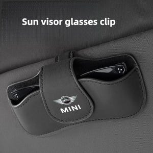 May include: A black leather sun visor glasses clip featuring the MINI logo and text. The holder is attached to a dark gray fabric sun visor and contains a pair of black-framed glasses. The text Sun visor glasses clip is visible above.