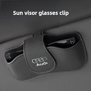 May include: A black leather sun visor glasses clip featuring the Audi logo in silver text on the front flap. The accessory is attached to a gray fabric car sun visor and contains a pair of black-framed glasses inside the storage compartment.