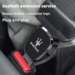 May include: A black Nappa leather seatbelt extension with a silver Maserati logo and text. The device features a metallic frame and is inserted into a standard red-button seatbelt buckle. Text on the image reads: Seatbelt extension service, Nappa leather + exclusive logo, Plug and play.