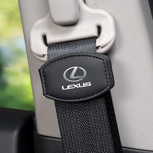 Pode incluir: Uma capa de cinto de segurança em couro preto com o logótipo e o texto Lexus em prateado. A capa está fixada numa correia de cinto cinzento-escuro, que passa por uma guia de plástico cinzento-claro montada no pilar interior do veículo.